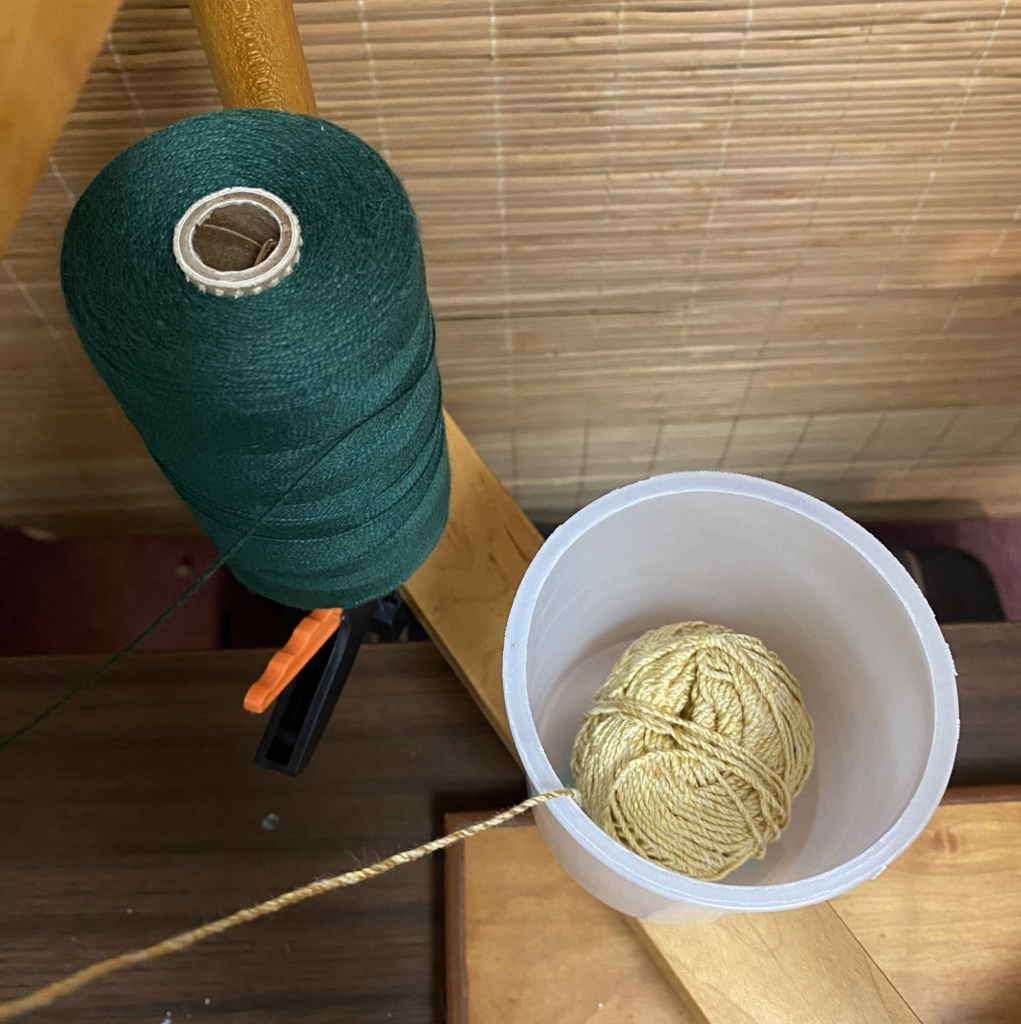 Photo of cone of yarn on a clamp with a ball of yarn in a cup next to it  on a warping mill.